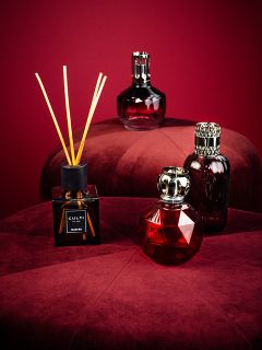 Vier elegante Parfümflakons, darunter ein Rohrdiffusor, sind auf zwei plüschigen, runden, weinroten Hockern vor einem passenden dunkelroten Hintergrund angeordnet. Die Flakons variieren in Form und Design und haben ein luxuriöses, anspruchsvolles Aussehen.