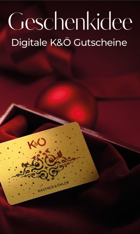 Eine goldene Kastner & Öhler Geschenkkarte ruht auf rotem Samt neben einem roten Ornament. Der Text lautet "Geschenkidee Digitale K&Ö Gutscheine" auf dunkelrotem Hintergrund.