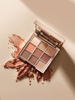 Eine quadratische Make-up-Palette mit neun Lidschatten in neutralen Tönen liegt offen auf einer beigen Fläche, auf der einige der pudrigen Farbtöne verstreut und zerdrückt sind. Im Deckel der Palette ist ein Spiegel zu sehen.