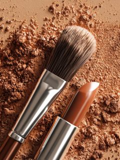 Eine Nahaufnahme eines Make-up-Pinsels und eines braunen Lippenstifts, die auf einer verstreuten Pudergrundierung in ähnlichen Brauntönen liegen, wodurch ein strukturierter und zusammenhängender Hintergrund zum Thema Schönheit entsteht.
