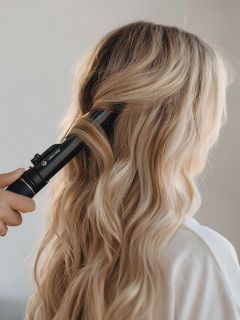 Eine Person mit langem, gewelltem blondem Haar wird von einer anderen Hand mit einem schwarzen Lockenstab gestylt. Der Fokus liegt auf dem Haar und dem Stylingprozess.