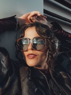 Eine Frau mit gewellten braunen Haaren trägt eine übergroße Sonnenbrille und eine dunkle Lederjacke. Sie lehnt an einer weißen Wand, die Hände über dem Kopf, und sieht entspannt und elegant aus.