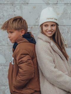 Ein Junge und ein Mädchen stehen Rücken an Rücken vor einer hellen Steinmauer. Der Junge trägt eine braune Jacke und blickt zur Seite, während das Mädchen einen wuscheligen Mantel und eine weiße Mütze trägt und lächelt, während es von dem Jungen wegschaut.