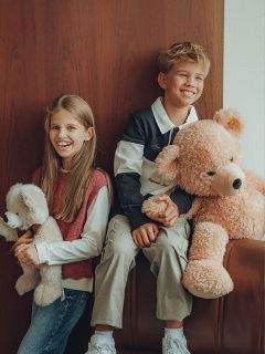 Zwei lächelnde Kinder sitzen nebeneinander und halten jeweils ein Stofftier. Das Mädchen hat lange Haare und umarmt einen kleineren Teddybären, während der Junge einen großen Teddybären hält. Sie sitzen an einer Holzwand und sehen glücklich und entspannt aus.