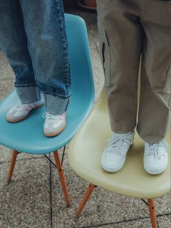 Zwei Kinder stehen auf Stühlen; eines auf einem blauen Stuhl und trägt Jeans und Turnschuhe mit rosa Streifen, das andere auf einem gelben Stuhl und trägt khakifarbene Hosen und weiße Turnschuhe. Nur ihre Beine und Füße sind zu sehen.