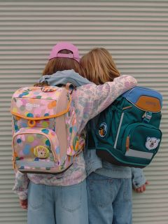 Zwei Kinder, die mit dem Rücken zur Kamera stehen, umarmen sich gegenseitig. Sie tragen bunte Rucksäcke - einer pastellfarben mit Mustern, der andere dunkelgrün mit Tiermotiven - und Freizeitkleidung, darunter Jeans und eine rosa Baseballmütze.