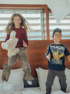 Zwei Kinder, ein Mädchen mit einem Stofftier in der Hand und ein Junge mit einem Pikachu-Shirt, hüpfen vergnügt auf einem Bett in einem hellen Raum mit großen Fenstern im Hintergrund.