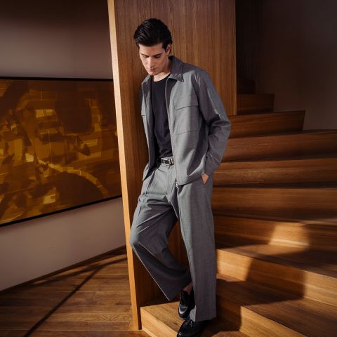 Ein Mann in einem grauen Anzug mit schwarzem Hemd und schwarzen Schuhen steht an einer Holztreppe und lehnt lässig an einer Wand in einem modernen Interieur.