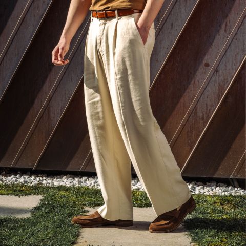 Eine Person steht im Freien und trägt eine cremefarbene Bundfaltenhose, einen braunen Gürtel, ein hellbraunes Hemd und braune Schuhe, wobei das Sonnenlicht Schatten auf den Boden wirft.