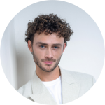 Mann mit lockigem braunem Haar und gestutztem Bart, der einen hellen Blazer und ein weißes Hemd trägt, steht vor einem weißen Hintergrund und lächelt sanft.