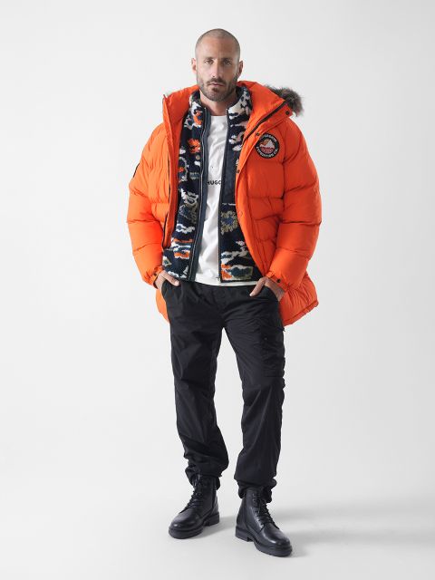 Ein Mann steht vor einem weißen Hintergrund und trägt eine leuchtend orangefarbene Pufferjacke mit einer pelzigen Kapuze, eine gemusterte Fleecejacke, ein weißes T-Shirt, eine schwarze Hose und schwarze Schnürstiefel.