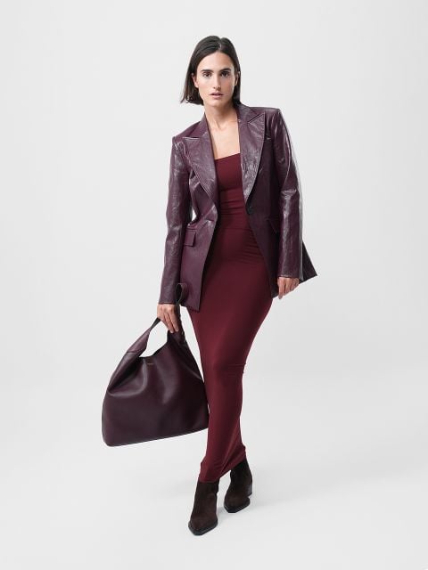 Eine Frau steht vor einem schlichten Hintergrund und trägt ein kastanienbraunes Kleid, einen passenden kastanienbraunen Lederblazer, braune Stiefeletten und eine große kastanienbraune Handtasche.