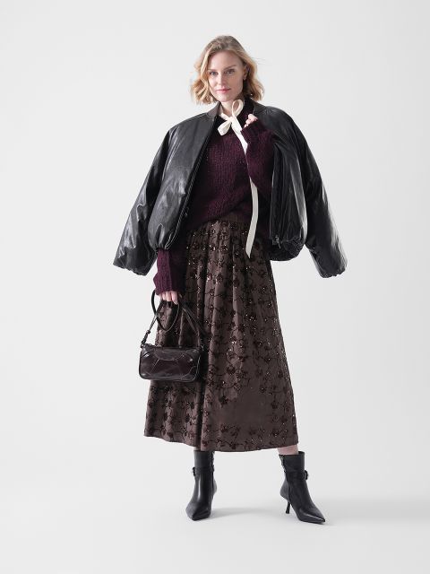 Eine Frau posiert in einer schwarzen übergroßen Lederjacke, einem kastanienbraunen Pullover und einem langen braunen geblümten Rock. Sie hält eine kleine Handtasche in der Hand und trägt schwarze Ankle Boots vor einem schlichten weißen Hintergrund.