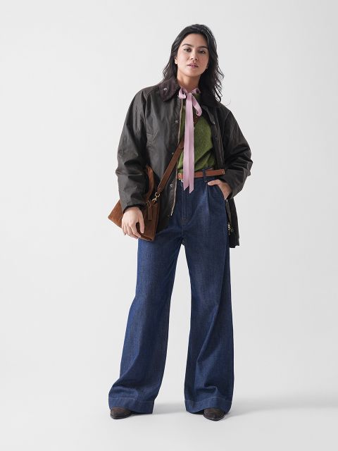 Eine Frau steht vor einem schlichten Hintergrund und trägt eine dunkelbraune Jacke, einen grünen Pullover mit einer rosa Schleife am Hals, eine weit geschnittene Bluejeans, braune Schuhe und eine braune Umhängetasche.