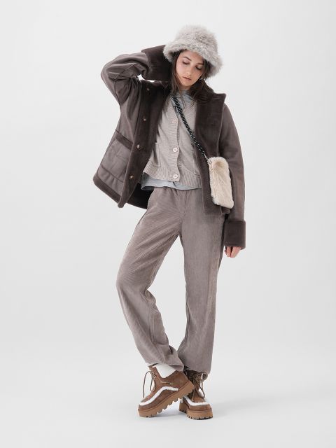 Eine Frau, die eine graue Pelzmütze, eine übergroße braune Jacke, einen hellgrauen Pullover, eine weit geschnittene Hose und braune Plateaustiefel trägt, posiert vor einem schlichten weißen Hintergrund. Sie hat eine kleine beigefarbene Pelztasche um ihren Körper geschlungen.