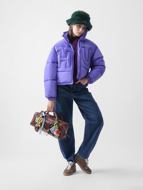 Eine Person posiert in einer lila Pufferjacke, blauen Jeans, Schuhen mit Leopardenmuster und einer grünen Pelzmütze. Sie hält eine braune Handtasche, die mit bunten Schlüsselanhängern und Bändern vor einem schlichten weißen Hintergrund verziert ist.