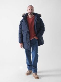 Ein Mann steht vor einem schlichten weißen Hintergrund und trägt eine marineblaue Pufferjacke mit einer pelzgefütterten Kapuze, einen roten Pullover, blaue Jeans und hellbraune Schuhe.