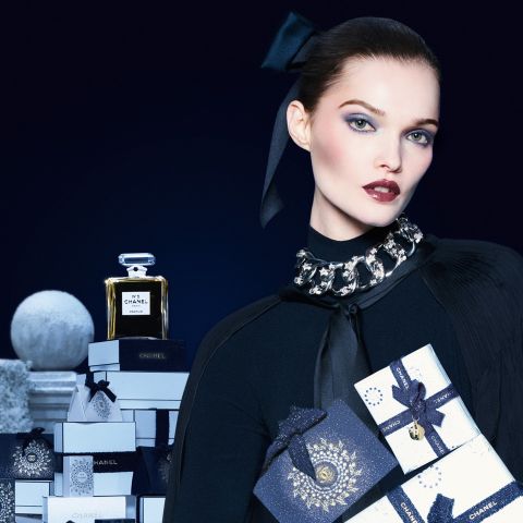Eine Frau in eleganter schwarzer Kleidung hält Chanel-Geschenkschachteln, daneben eine Flasche Chanel No. 5-Parfüm, vor einem dunkelblauen Hintergrund.
