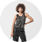 Eine Frau mit lockigem Haar posiert in einem ärmellosen schwarzen Paillettenoberteil und einer schwarzen Lederhose, in der Hand eine kleine Handtasche, vor einem schlichten hellen Hintergrund.