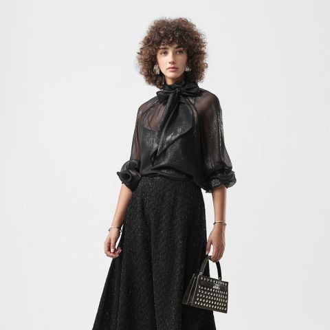 Frau mit lockigem Haar in schwarzer, durchsichtiger Bluse mit Schleife und langem, strukturiertem schwarzem Rock, die eine mit Nieten besetzte Handtasche in der Hand hält, steht vor einem schlichten Hintergrund.