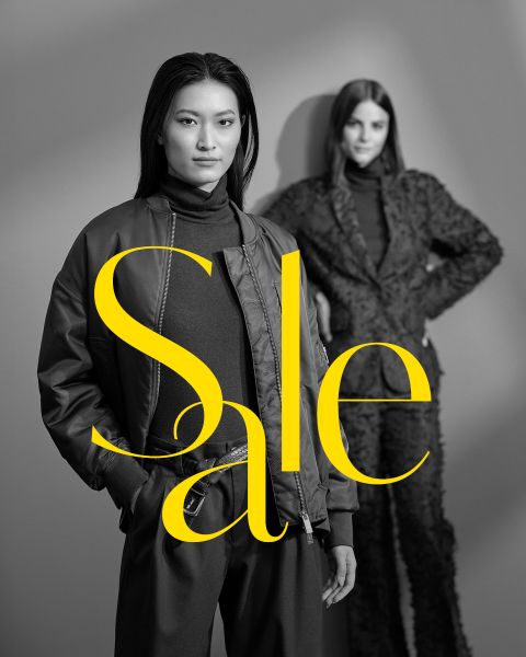 Zwei Frauen posieren in stilvollen dunklen Outfits vor einem grauen Hintergrund mit dem Wort "Sale" in großen gelben Buchstaben quer über dem Bild.