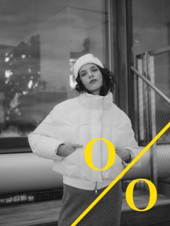 Eine Person in weißer Pufferjacke und Mütze posiert im Freien in der Nähe eines Gebäudes; eine gelbe diagonale Linie durchquert das Bild mit zwei gelben "O"-Buchstaben auf beiden Seiten. Das Foto ist in schwarz-weiß gehalten.