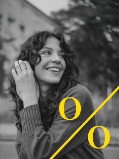 Eine Frau mit lockigem Haar lächelt und blickt zur Seite. Sie trägt einen Pullover und sitzt im Freien. Ein großes gelbes Prozentzeichen überlagert das Schwarz-Weiß-Foto.