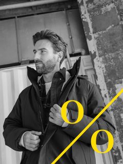 Ein Mann mit Bart steht vor einem industriellen Hintergrund und rückt seine Jacke zurecht. Das Schwarz-Weiß-Foto zeigt fette gelbe Prozentzeichen und eine diagonale gelbe Linie in der rechten unteren Ecke.