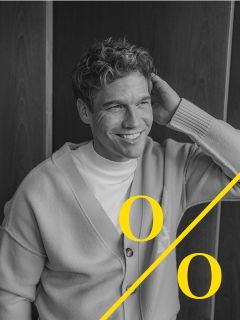 Ein Mann mit lockigem Haar, der eine helle Strickjacke und ein weißes Hemd trägt, sitzt lächelnd da und hält eine Hand hinter den Kopf. Das Schwarz-Weiß-Bild zeigt zwei große gelbe "o "s und eine diagonale gelbe Linie am unteren Rand.