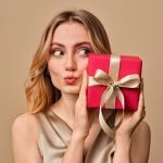 Frau mit blondem Haar hält eine rote Geschenkbox mit goldener Schleife und macht ein verspieltes Kussmundgesicht vor einem beigen Hintergrund.