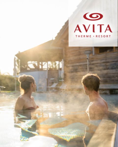 Eine Frau und ein Mann entspannen sich in einem dampfenden Außenpool im AVITA Therme Resort, mit einem Holzgebäude und sanftem Sonnenlicht im Hintergrund.