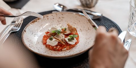 Eine Person ist im Begriff, ein Gericht mit gehackten Tomaten, Mozzarella, Basilikum und Pesto auf einem rustikalen Teller zu essen, der auf einem schwarzen Tischtuch steht.