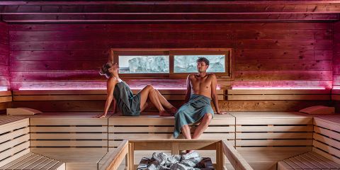 Zwei Personen in Handtüchern entspannen sich auf Holzbänken in einer modernen Sauna mit sanfter Beleuchtung und einem kleinen Fenster, das den Blick auf die Felsen draußen freigibt.