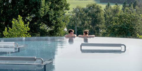 Zwei Personen entspannen sich in einem Infinity-Pool im Freien, umgeben von Bäumen und mit Blick auf eine üppige, grüne Landschaft.