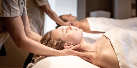 Zwei Personen erhalten eine entspannende Nackenmassage, während sie auf Massagetischen liegen, die jeweils mit weißen Handtüchern bedeckt sind, in einer beruhigenden Spa-Umgebung.