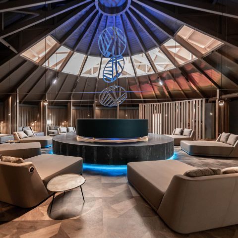 Moderne Lounge mit kreisförmigen Sitzgelegenheiten, weichen beigen Sofas, einer runden zentralen Leuchte und einem geometrischen Deckendekor unter einem hohen, gewölbten Holzdach mit blauer Beleuchtung.