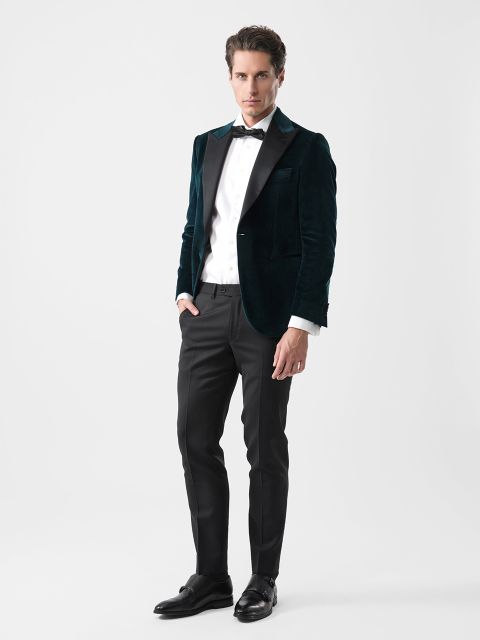 Ein Mann in einem dunkelgrünen Samtblazer, einem weißen Hemd, einer schwarzen Fliege und einer schwarzen Hose steht vor einem schlichten weißen Hintergrund.