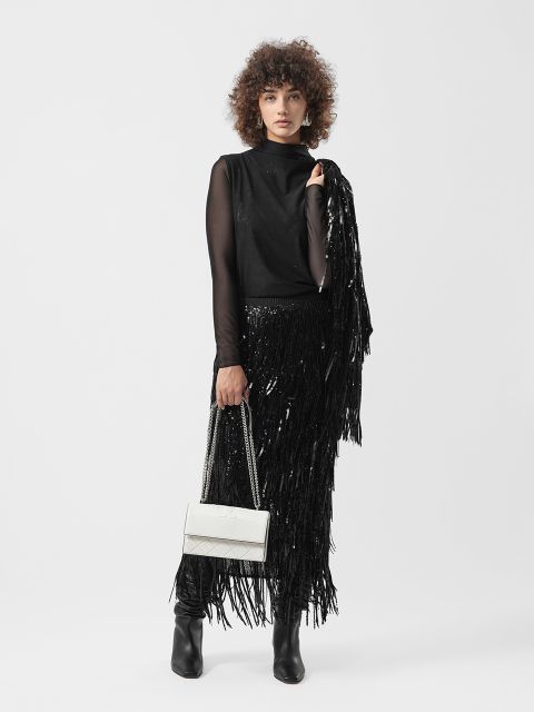 Frau mit lockigem Haar in schwarzem Fransenrock und Oberteil, mit weißer Handtasche, in schwarzen Stiefeln, vor einem schlichten weißen Hintergrund.