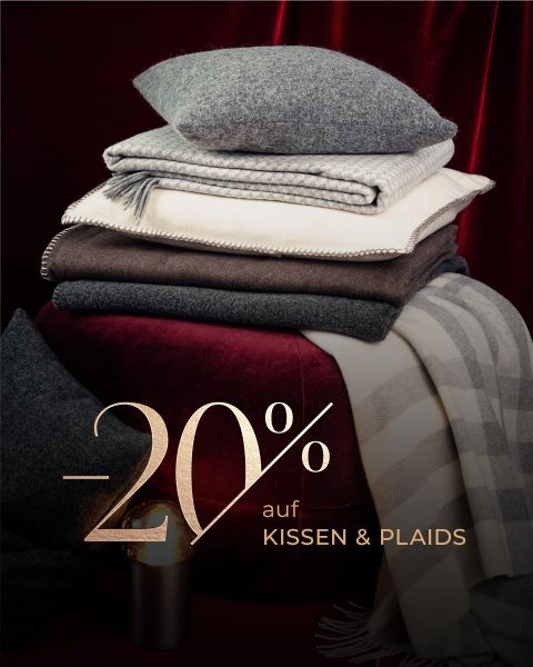 Ein Stapel gefalteter Kissen und Decken auf einer roten Samtfläche mit Text: "-20% auf KISSEN & PLAIDS" in goldener Schrift.
