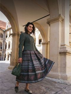 Eine Frau in einem gemusterten Rock und einer grünen Jacke steht unter steinernen Bögen, lächelt und hält eine grüne Handtasche in der Hand.