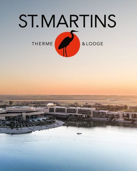 Luftaufnahme der St. Martins Therme & Lodge an einem See bei Sonnenuntergang, mit dem Logo und einer Vogelgrafik über den Gebäuden des Resorts.