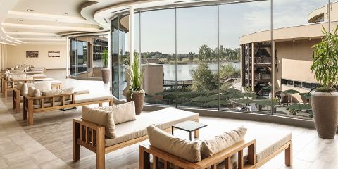 Moderner Lounge-Bereich mit gepolsterten Holzsitzen, großen Topfpflanzen und bodentiefen Fenstern mit Blick auf einen See und Außengebäude.