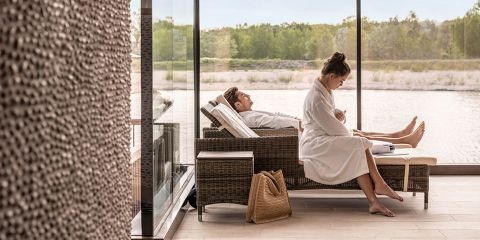 Zwei Personen in weißen Bademänteln entspannen sich auf Liegestühlen an großen Fenstern mit Blick auf einen ruhigen See und Bäume, während eine gewebte Tasche auf dem Boden liegt.