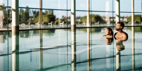 Ein Mann und eine Frau entspannen sich in einem großen Hallenbad mit bodentiefen Fenstern und genießen das Sonnenlicht und die Aussicht auf die grüne Umgebung.