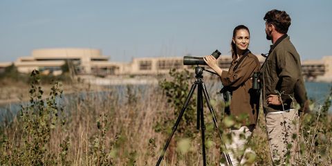 Zwei Personen, die mit einem Spektiv auf einem Stativ Vögel in der Nähe von Gewässern beobachten, mit Gebäuden und Grünflächen im Hintergrund unter einem klaren Himmel.