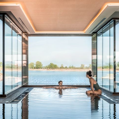 Zwei Personen entspannen sich in einem Innenpool mit raumhohen Glaswänden und Blick auf einen malerischen See und Bäume unter einem klaren Himmel.