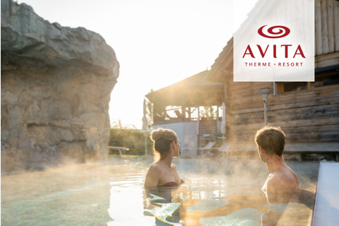 Zwei Personen entspannen sich in einem dampfenden Außenpool in einem Kurort mit Holzgebäuden und Felsen in der Nähe. In der Ecke ist das Logo des AVITA Therme Resorts zu sehen.