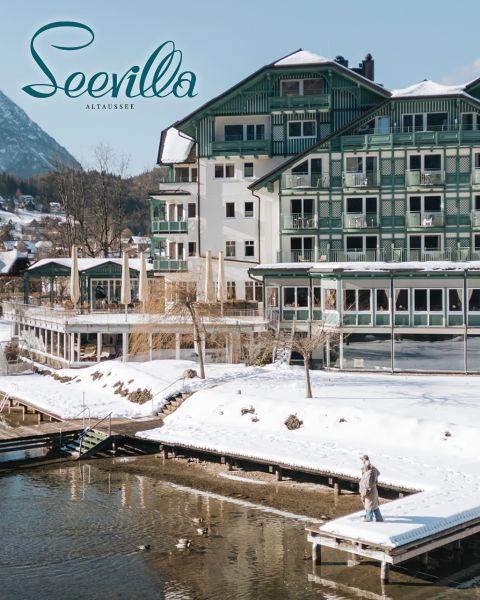 Ein Hotel am See mit grünen Balkonen und verschneiten Dächern, Berge im Hintergrund und eine Person, die auf einem Steg am Wasser steht.