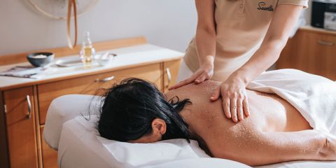 Eine Person erhält eine Rückenmassage mit Peeling in einem Wellnessbereich; der Therapeut wendet die Behandlung an, während der Kunde auf einer Massageliege liegt.