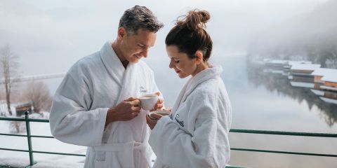 Ein Paar in weißen Bademänteln genießt ein heißes Getränk auf einem Balkon mit Blick auf einen ruhigen See und eine schneebedeckte Landschaft.
